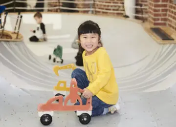 young girl in yellow shirt smiling and crouched in play area bowl with car on wheels