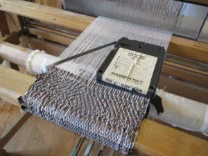 A close up image of a floor loom with white string as the warp and 8-track tape as the weft. An 8-track tape sits on top of the woven fabric.
