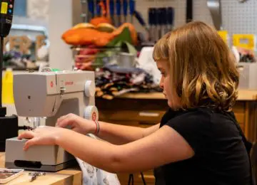 A child is feeding fabric through a sewing machine. She has one hand on either side of the sewing machine's needle as it stitches the fabric.