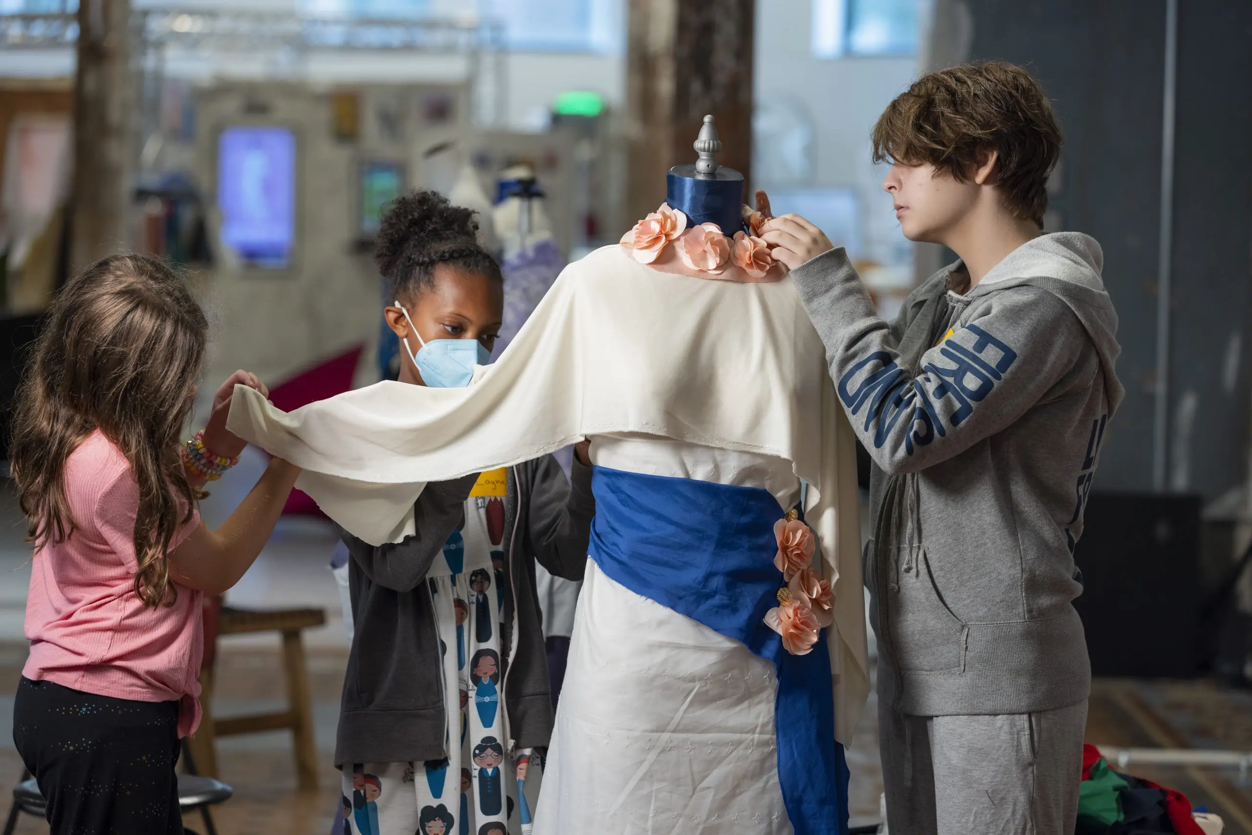 Three kids work on pinning fabric to a dress form