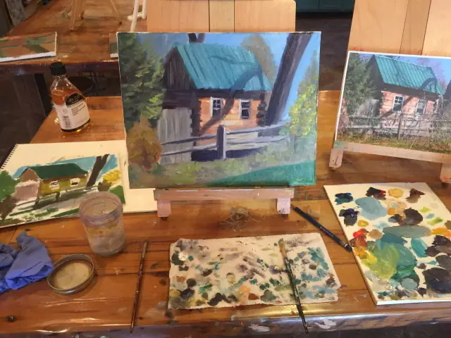 An oil painting of a house sits on a tabletop easel. On the table around the painting are painting materials such as a palette, brushes, a water cup, and a reference photo.