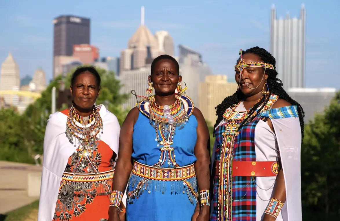 Three women wearing beaded jewelry stand in front of Pittsburgh's skyline