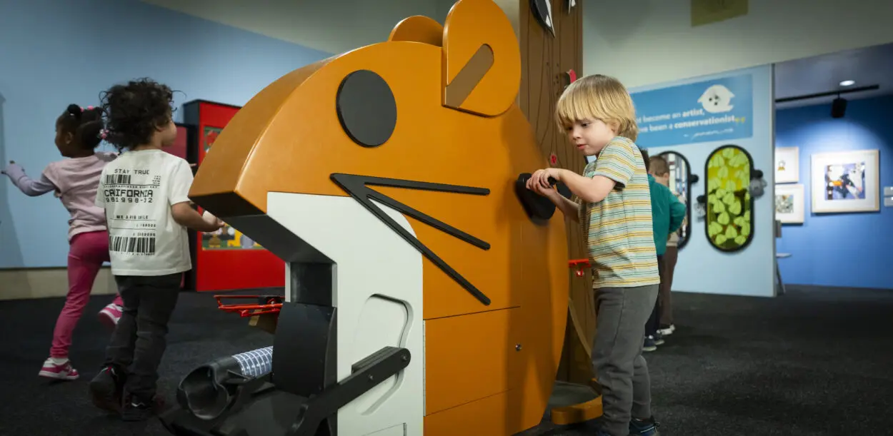 young children play around a larger-than-life mouse feature and one rotates a hand crank
