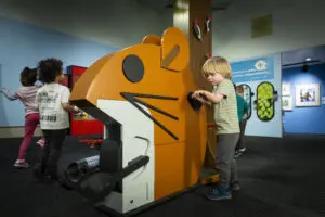 young children play around a larger-than-life mouse feature and one rotates a hand crank