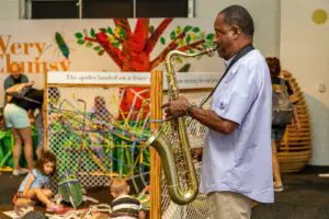 Man playing saxophone in a room where kids are building and weaving on the floor, in an Eric Carle exhibit