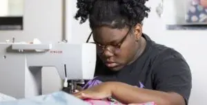 A young, Black learner with glasses guides fabric through a sewing machine