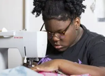 A young, Black learner with glasses guides fabric through a sewing machine