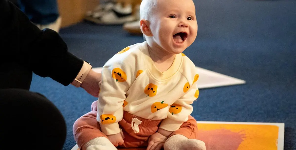 Infant in pumpkin shirt and orange pants sitting on the floor and laughing