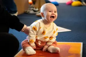 Infant in pumpkin shirt and orange pants sitting on the floor and laughing