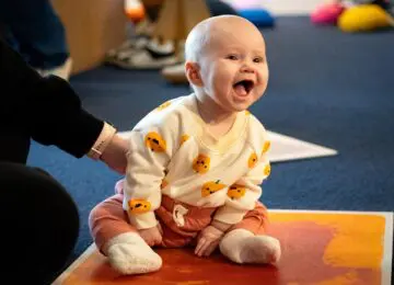 Infant in pumpkin shirt and orange pants sitting on the floor and laughing