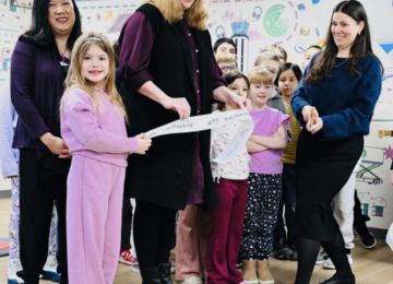 3 women and several children cut a ribbon in a colorful room and laugh