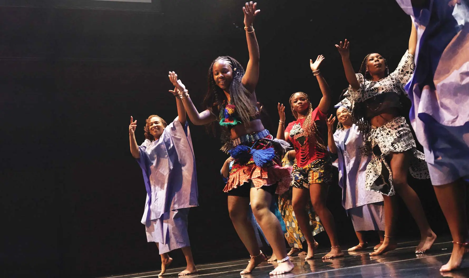 Several women dancing on a stage in traditional African clothing with arms raised up