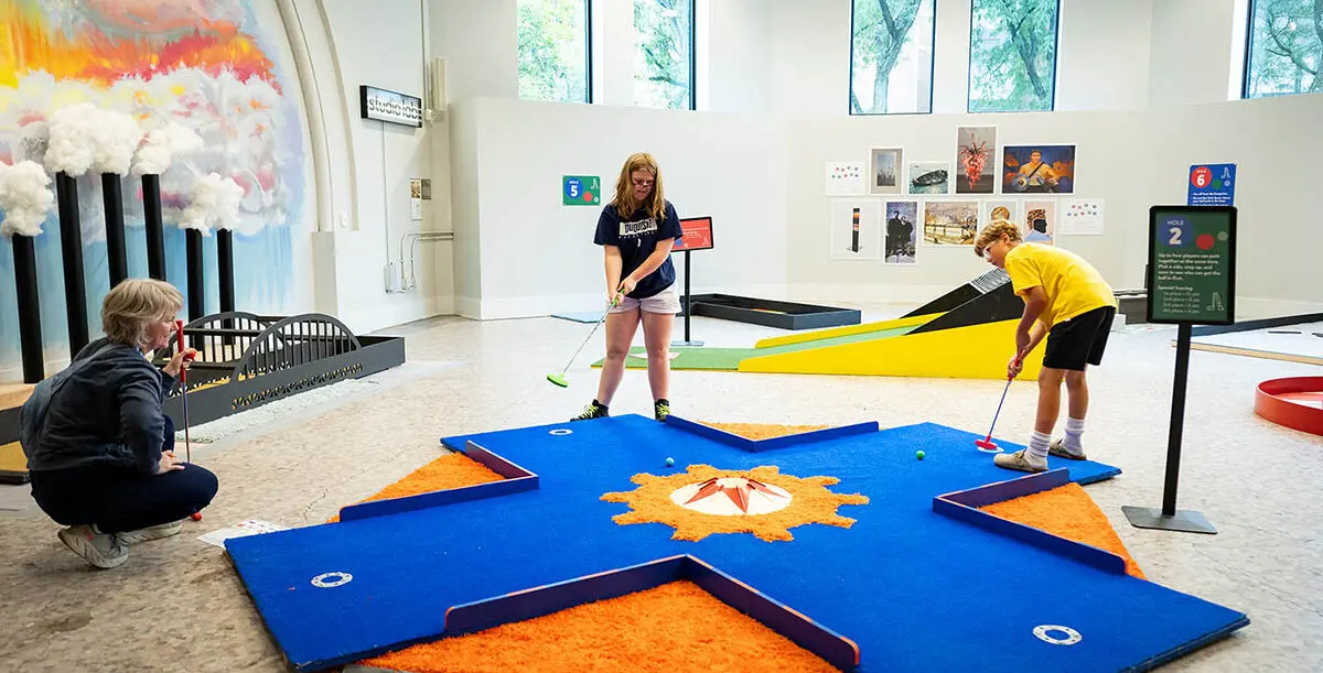 three people playing min golf indoors, two youth and an adult, in a white room with large windows