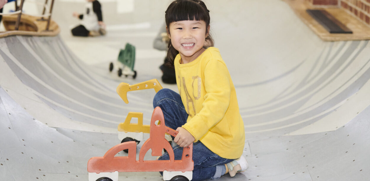 young girl in yellow shirt smiling and crouched in play area bowl with car on wheels
