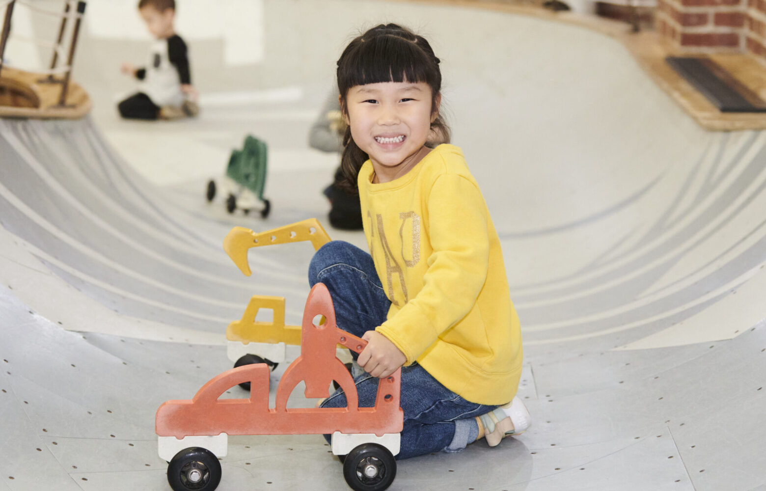 young girl in yellow shirt smiling and crouched in play area bowl with car on wheels