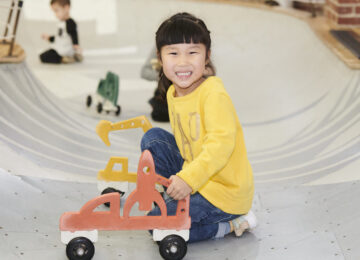 young girl in yellow shirt smiling and crouched in play area bowl with car on wheels