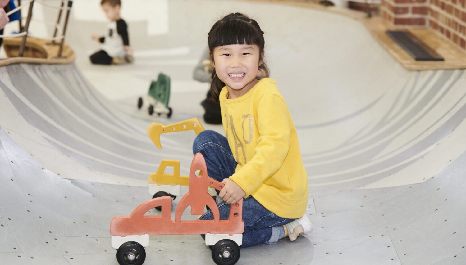 young girl in yellow shirt smiling and crouched in play area bowl with car on wheels