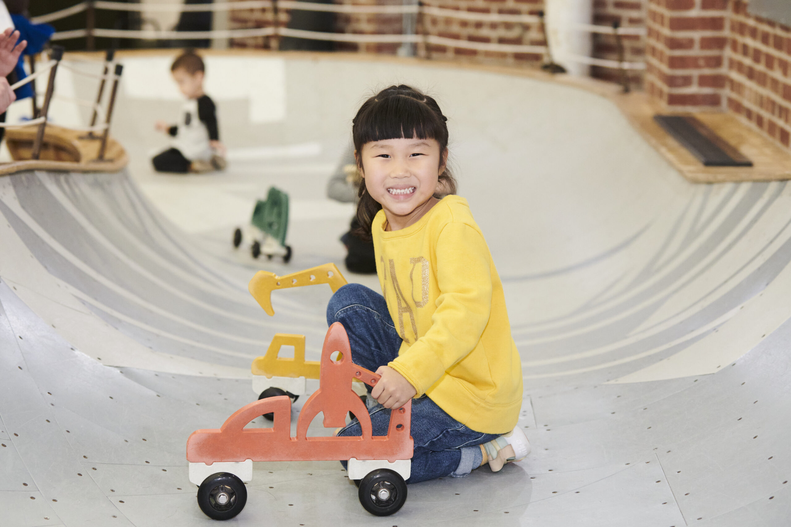 young girl in yellow shirt smiling and crouched in play area bowl with car on wheels