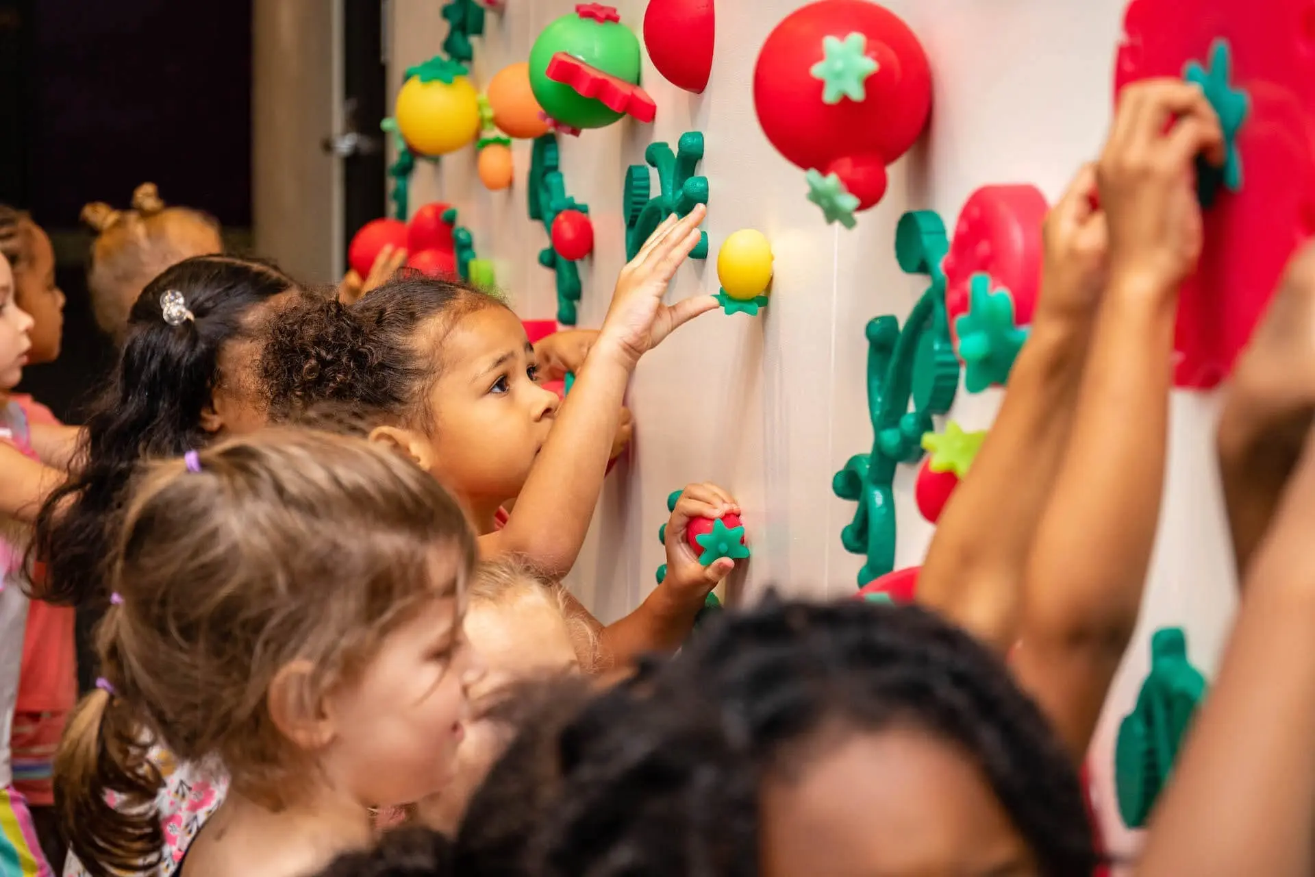 group of young children touching silicone objects in the shape of fruits and vegetables mounted on a wall