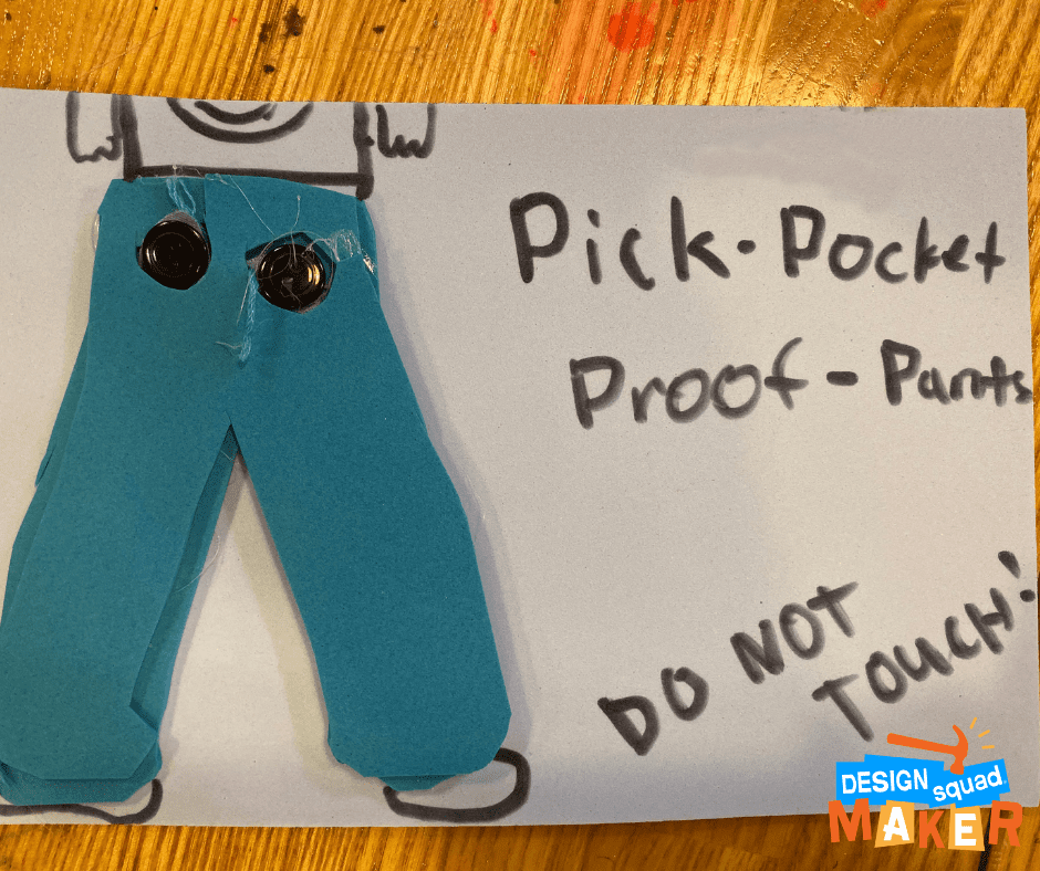 A paper with the words "pick-pocket proof-pants, do not touch." Additionally on the paper, there is blue foam in the shape of pants with two clothes buttons attached to it.
