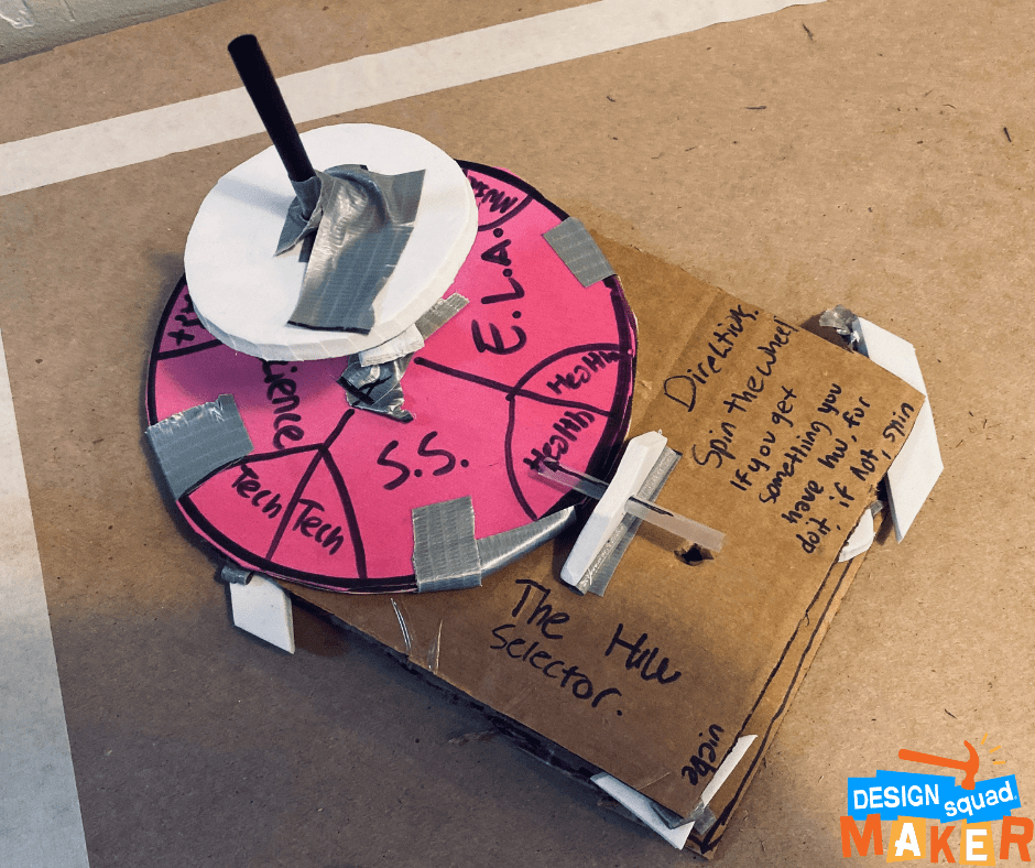 A cardboard box with a pink circular wheel above it, followed by a white wheel above the pink wheel. The brown box has writing on it saying: The homework selector. It also says: Directions-- Spin the wheel if you get something you have for homework, do it, if not, spin. The pink wheel is segmented with different school subjects from Tech, social studies, health, and E.L.A. There is a pointer made out of a sipping straw which points at one of the subjects on the wheel: health.