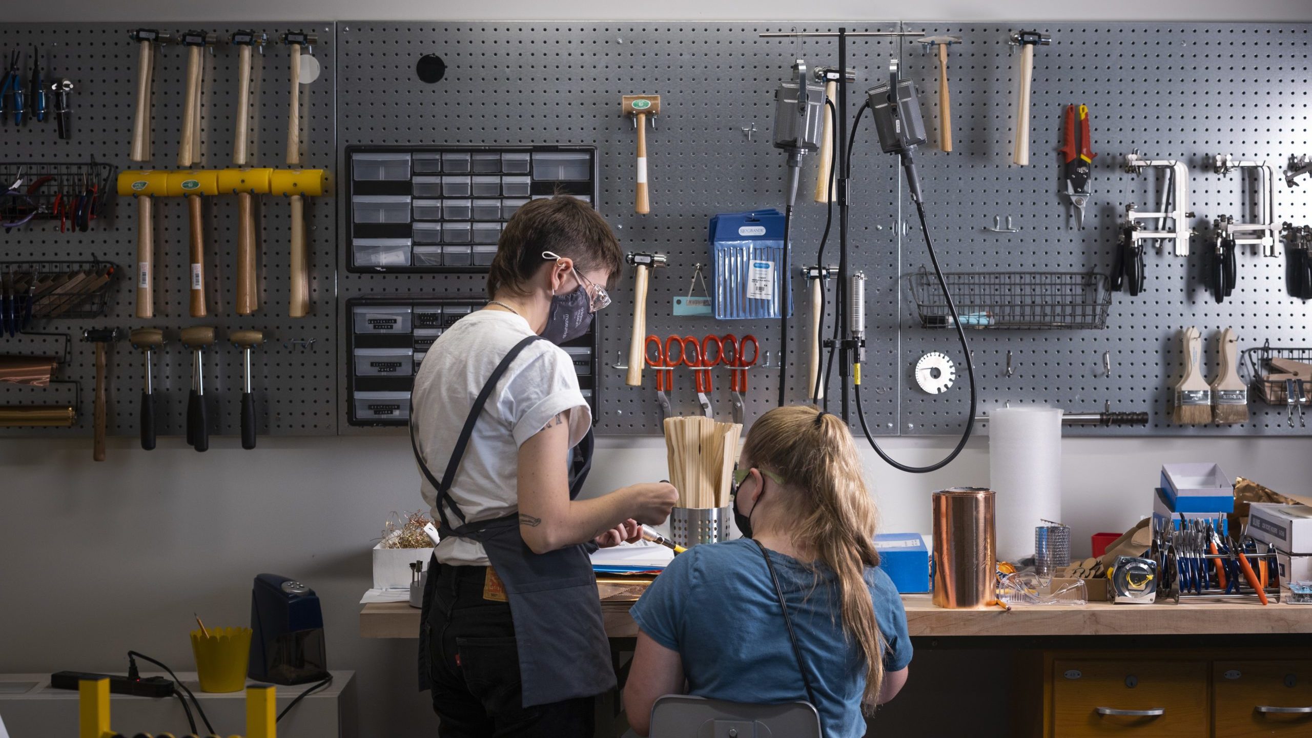 A photograph taken from behind of two people, an adult and a child. They are working together in front of a wall of metalworking tools.