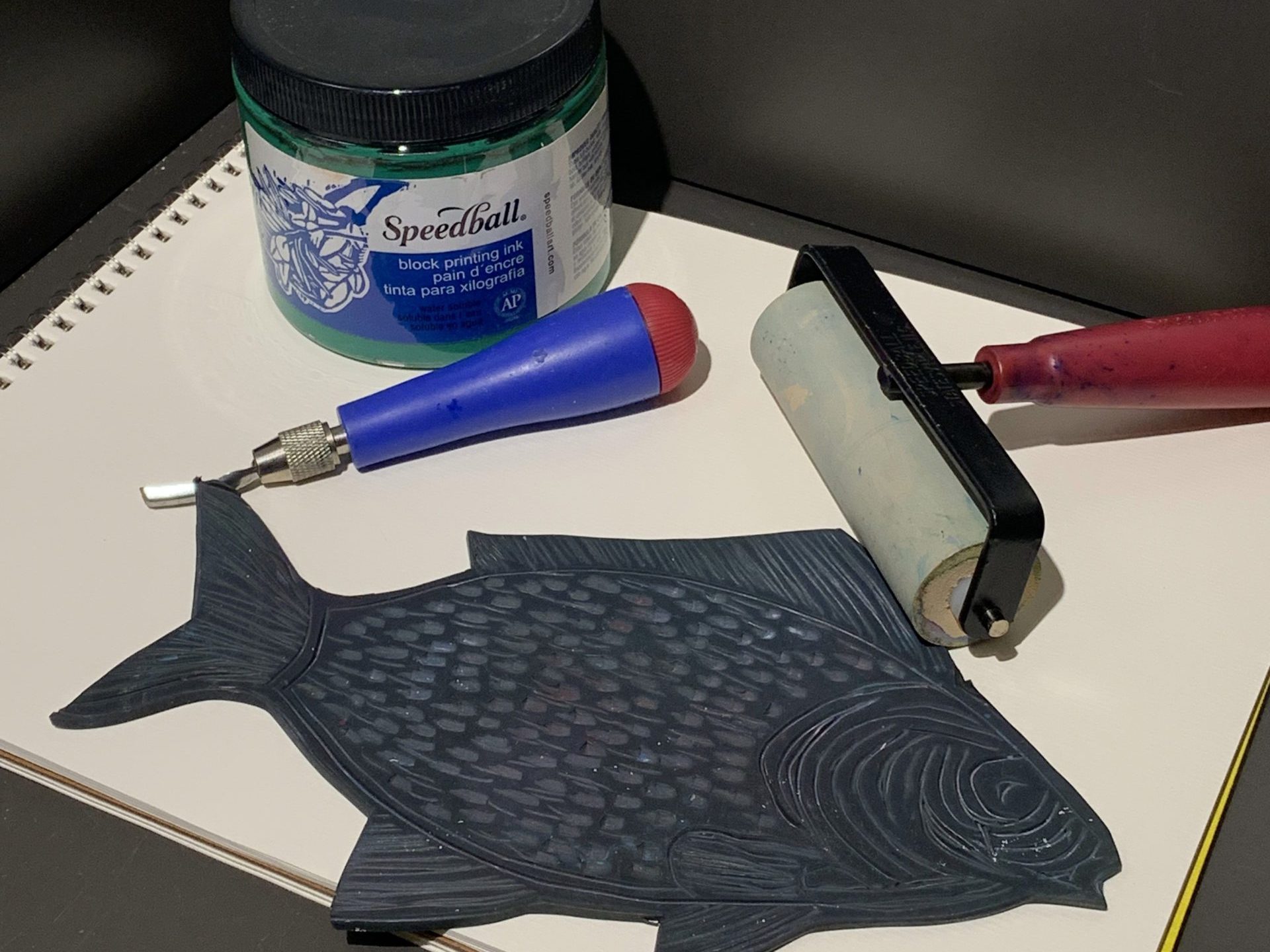 A tableau of printmaking items, including paper, a brayer, ink, a cutting tool, and a piece of linoleum carved to look like a fish.