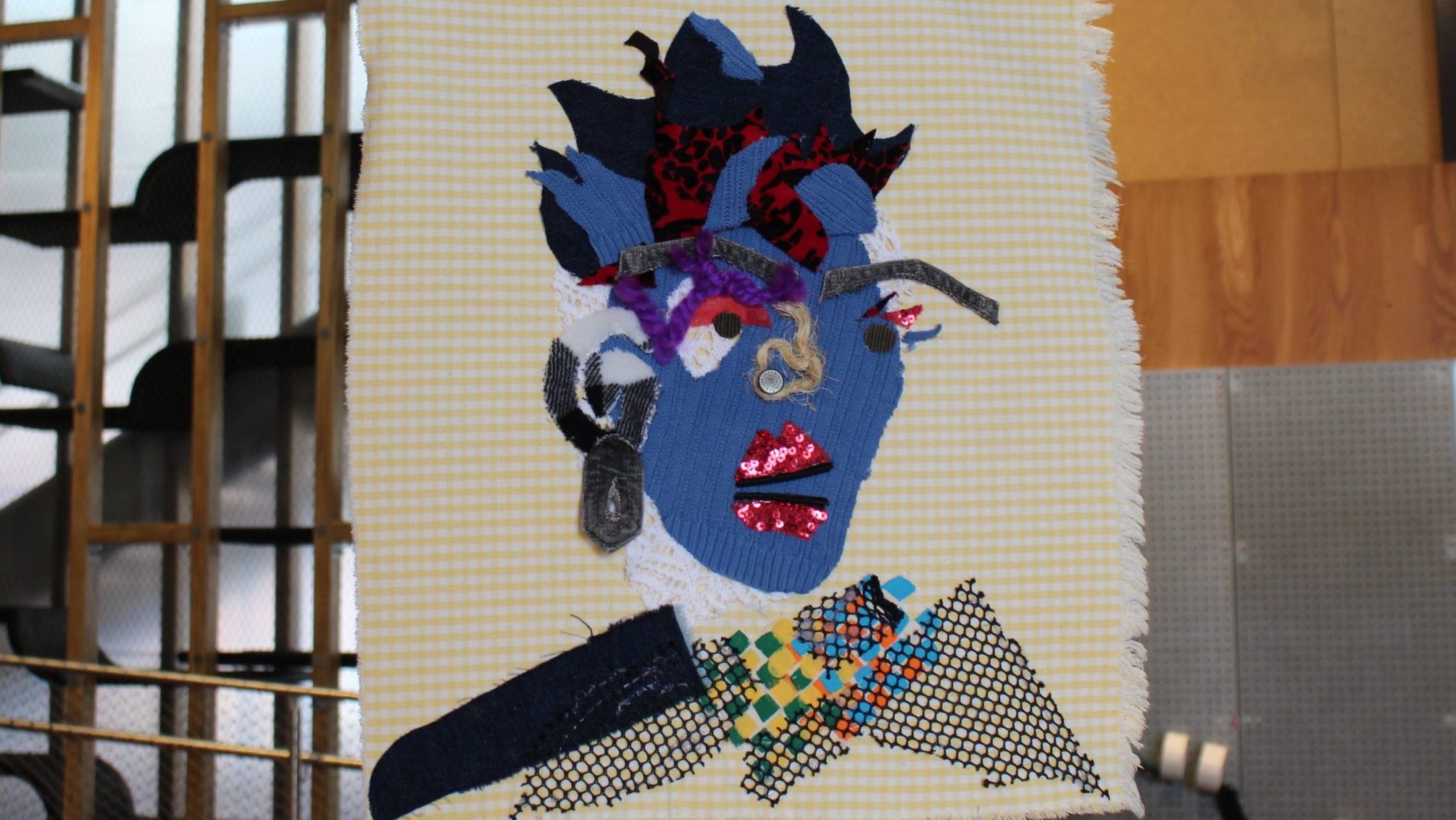 A colorful fabric collage of a face with blue skin, wild hair, shining lips and many visual and physical textures
