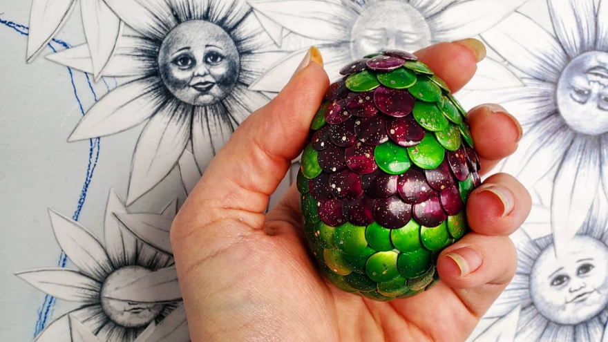 A hand with clean, longer length nails holds a green-and-purple metallic egg. It is a photo taken in front of a wall illustrated with smiling flowers.
