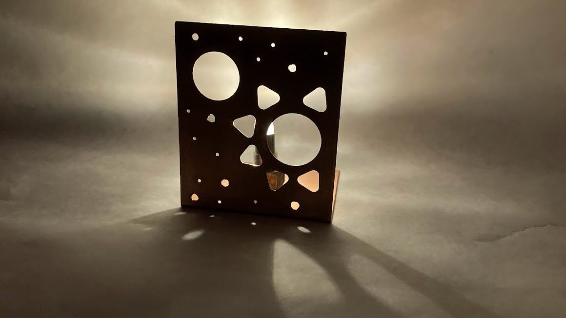 Light shines through a cut copper sheet, with rays and shadows emanating from the round and triangular holes.