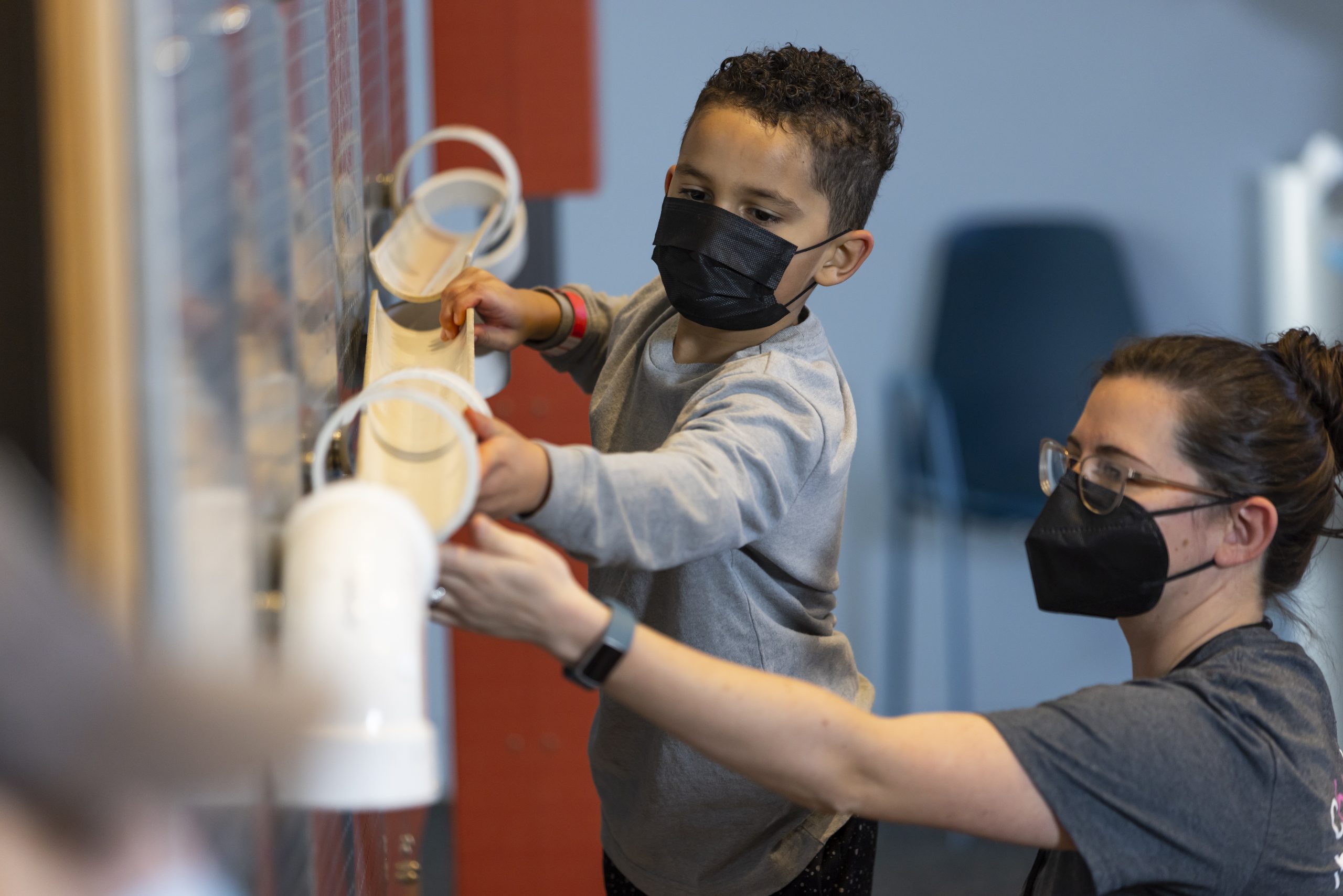A young visitor and a staff member, both wearing masks, are playing with the ball wall in MAKESHOP.