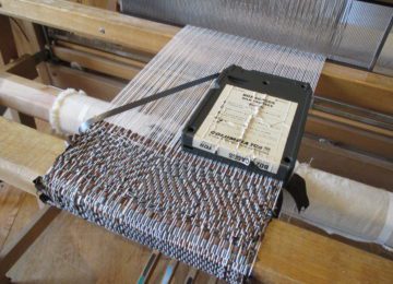 A close up image of a floor loom with white string as the warp and 8-track tape as the weft. An 8-track tape sits on top of the woven fabric.