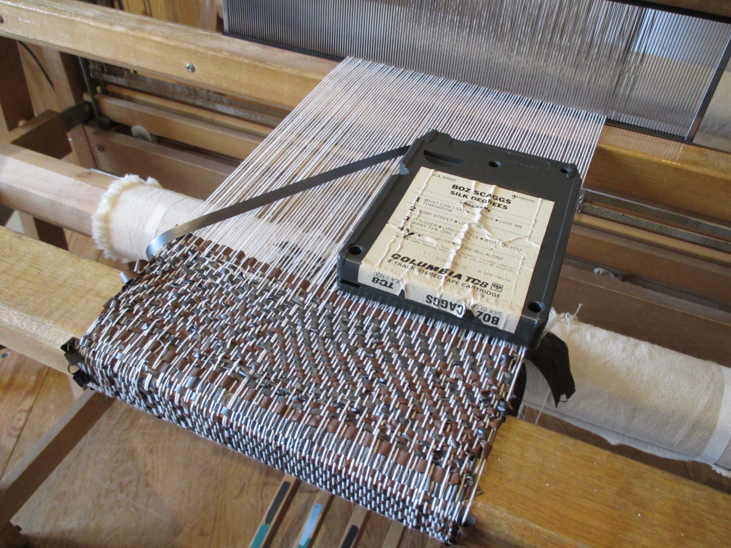 A close up image of a floor loom with white string as the warp and 8-track tape as the weft. An 8-track tape sits on top of the woven fabric.