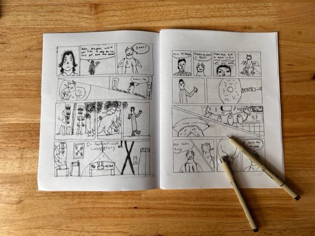 A graphic novel is laid open to reveal two pages of black-and-white illustrations. Two pens lay over the corner of the graphic novel.