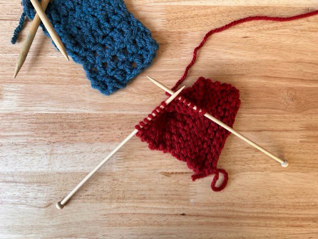 Two wooden knitting needles are attached to an in-progress knitted item, made from red yarn. A finished piece of knitting made from blue yarn is partially in frame in the top left.