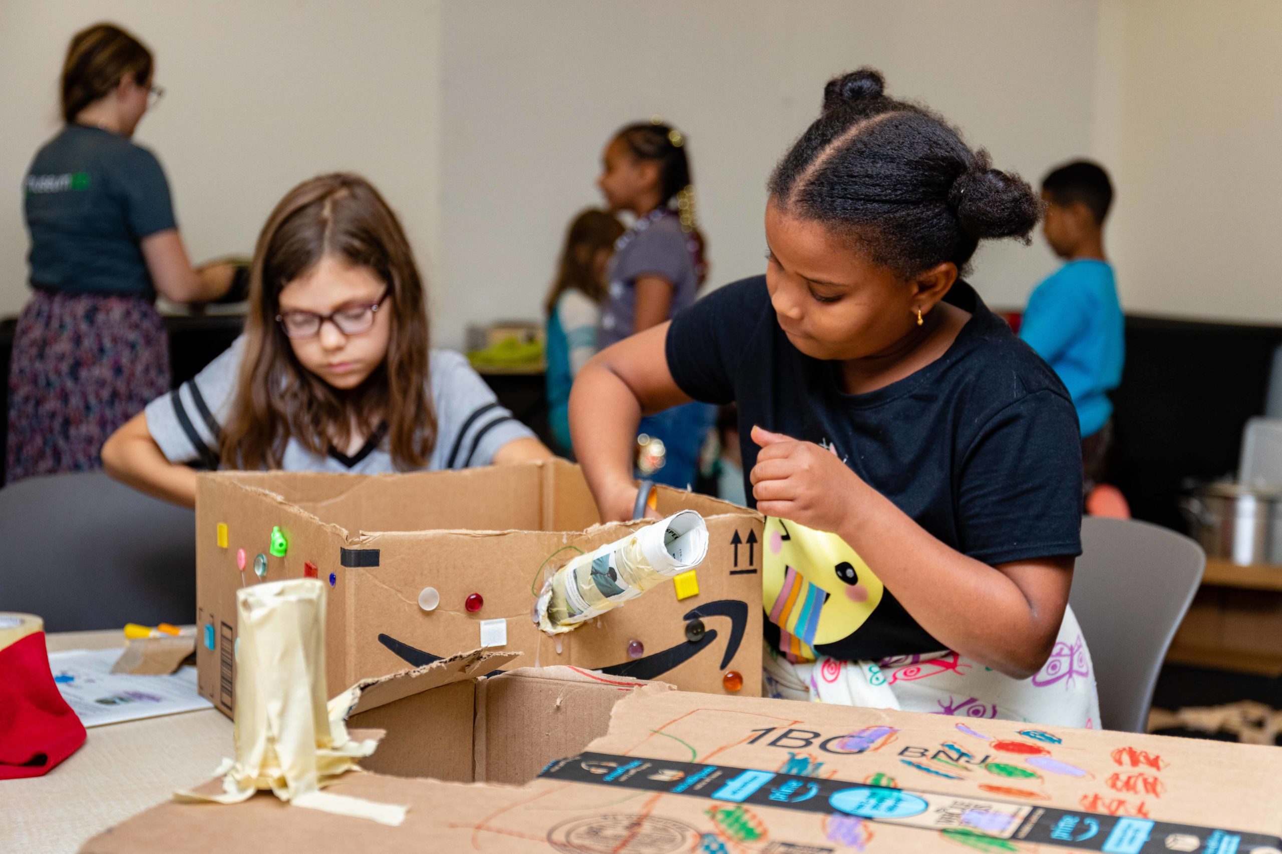 A child works on a creation made of a box, cardboard tubes, buttons, and other familiar materials. There are other children and a museum educator in the background working on their projects.