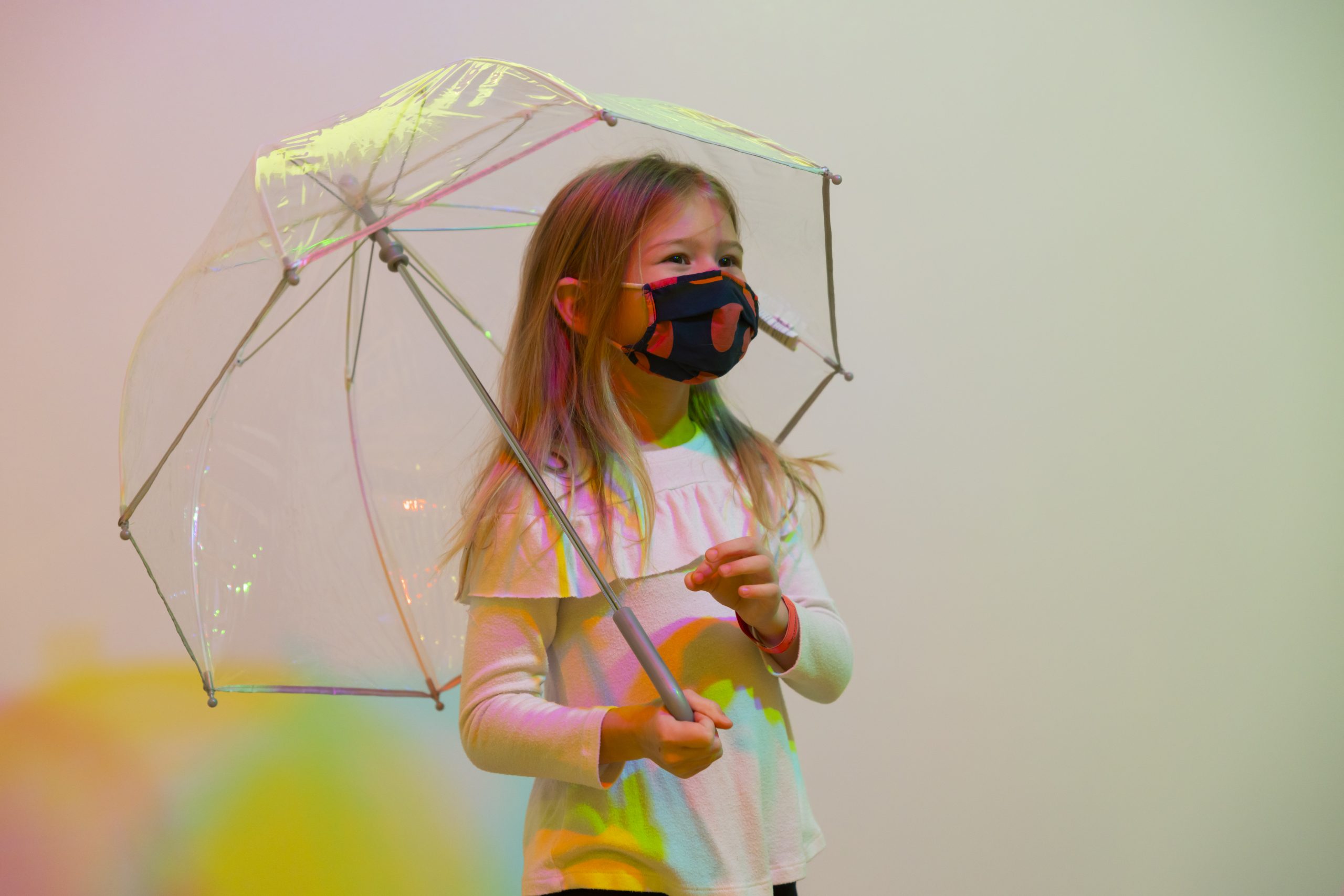 A young child holds a clear umbrella over her head. Red, yellow, and blue shadows are created behind her by lights (not pictured) hanging from above.