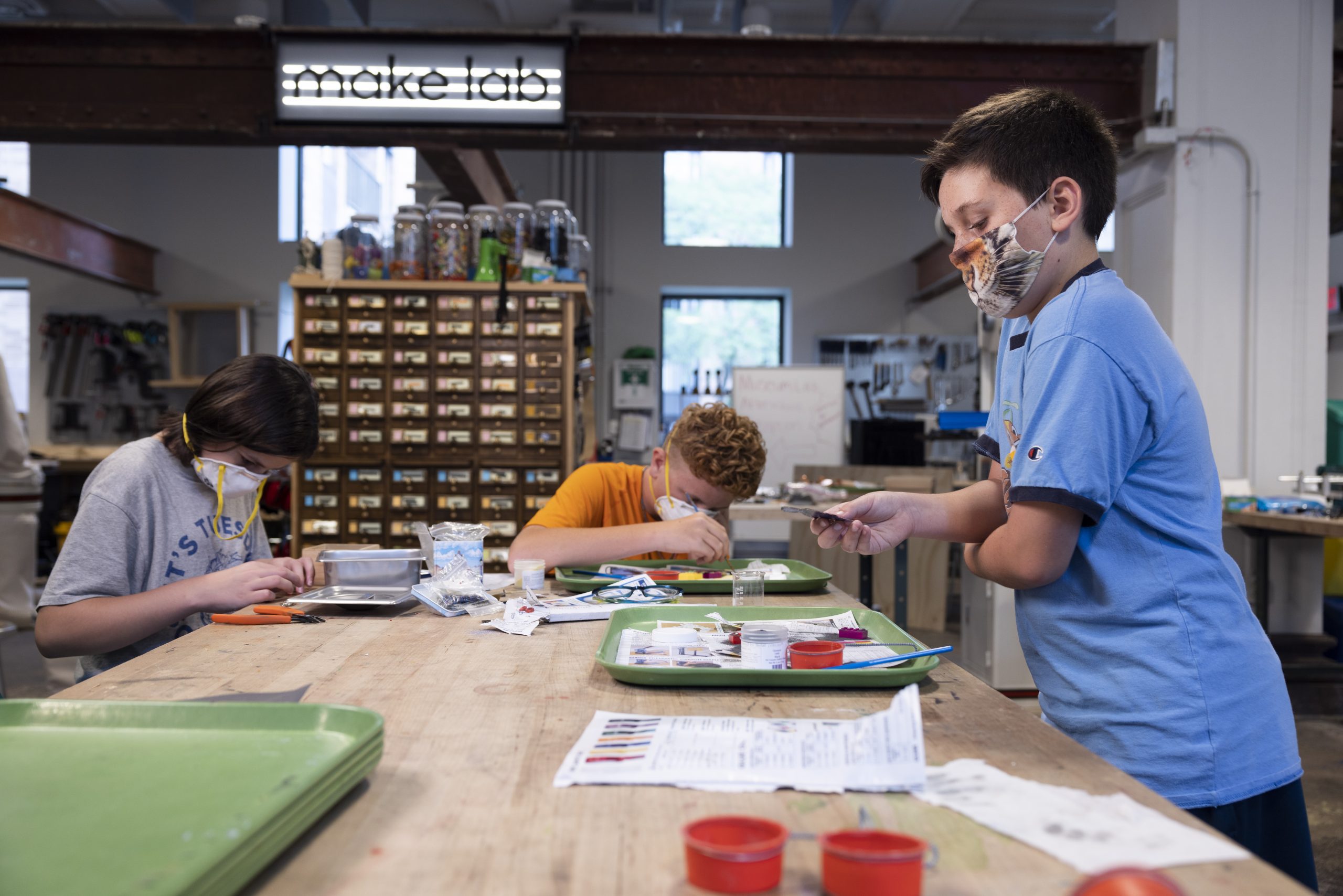 Three young people are working around a table. They are using glass enameling dust and sifters on top of green trays lined with newspaper.