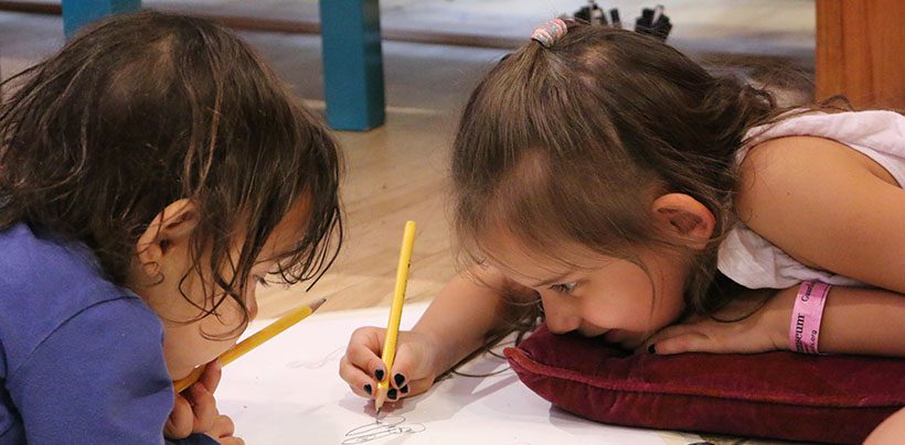 Two young children lean over a piece of paper. One child writes on the paper with a pencil.