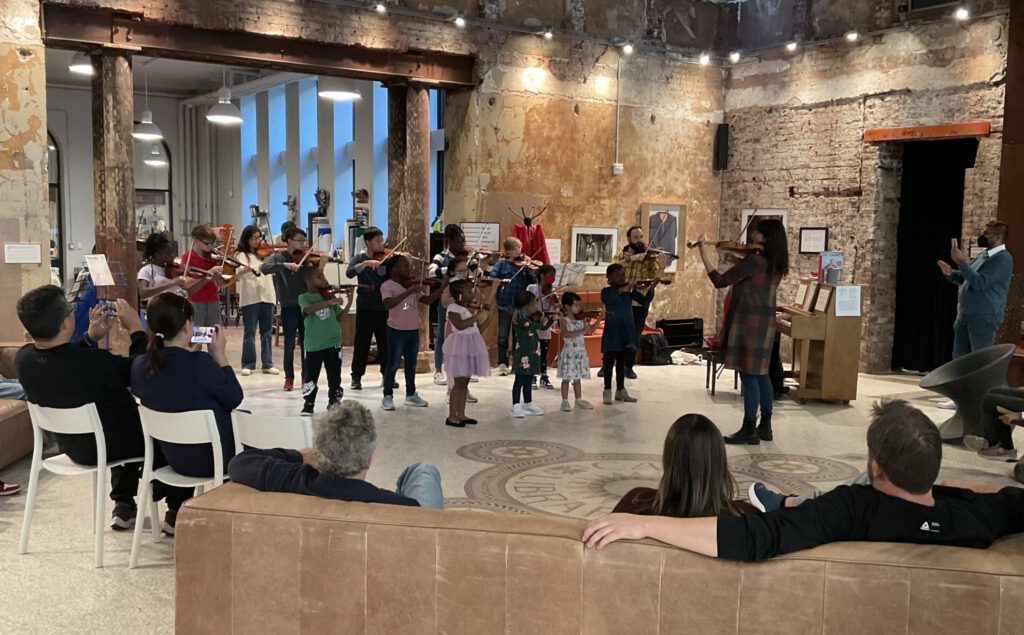 Children of various ages playing string instruments in concert.