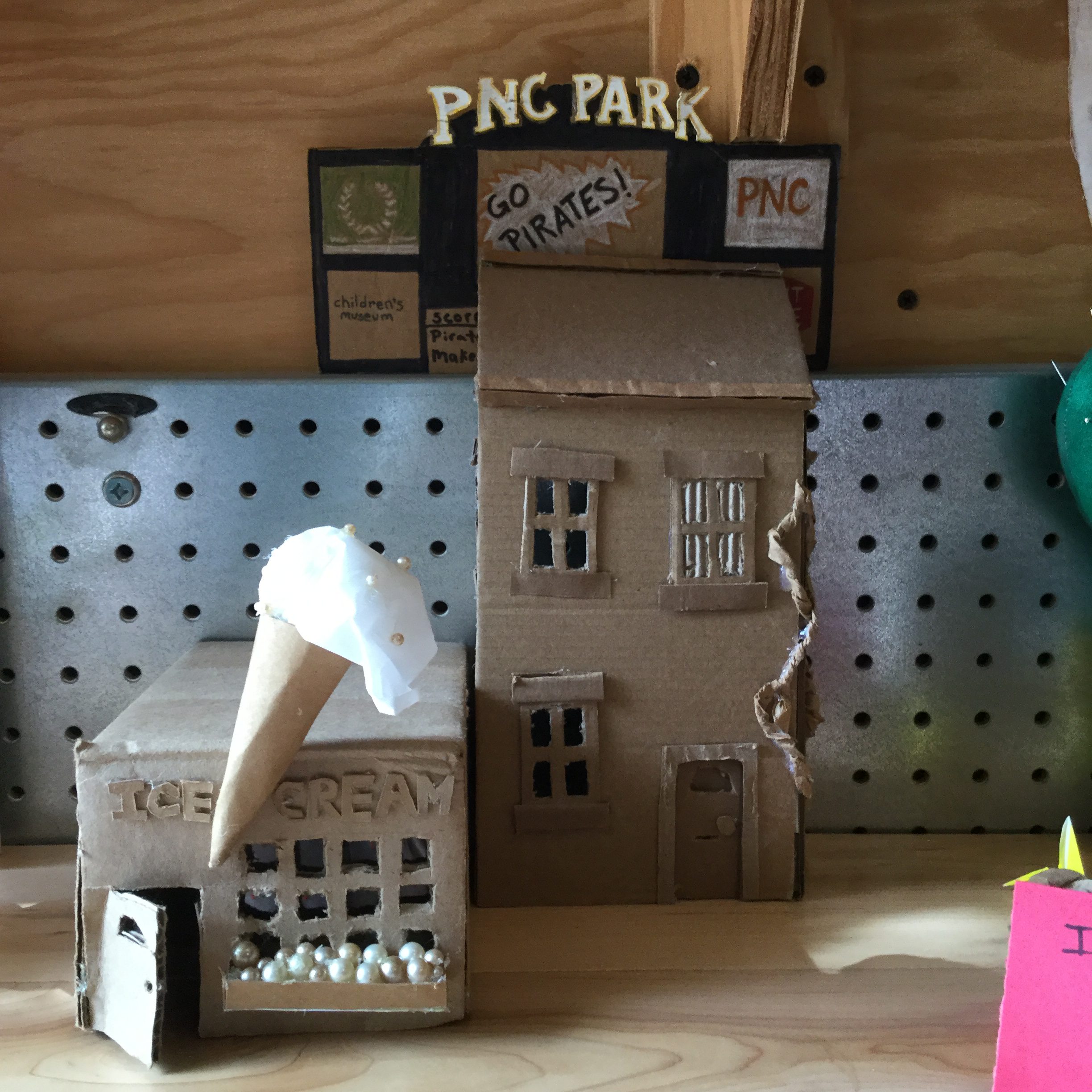 Two small-scale building made of cardboard and other recycled materials sit on a table. One is an ice cream shop and the other is a house.