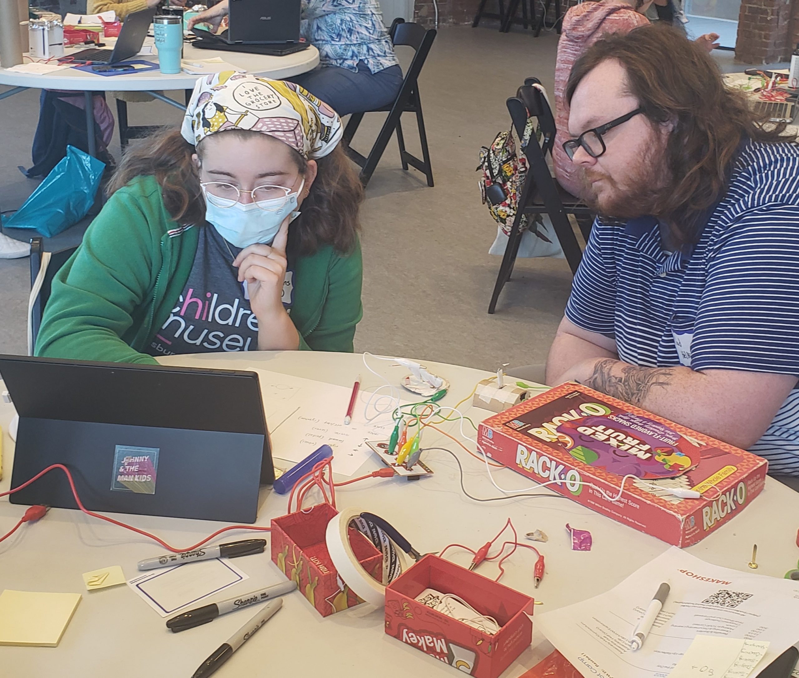 Two adults sit at a table looking intently at a laptop computer that is connected to a Makey Makey kit.