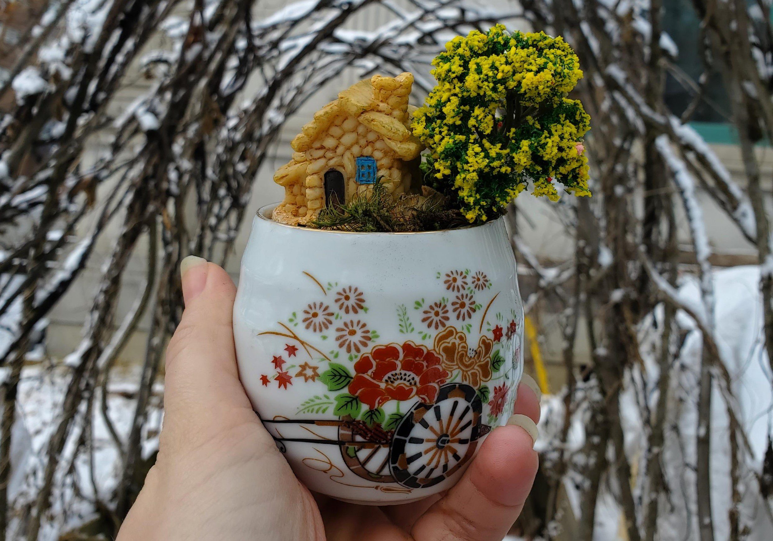 A hand holds up a small, porcelain teacup with a plant and a small, decorative house inside.