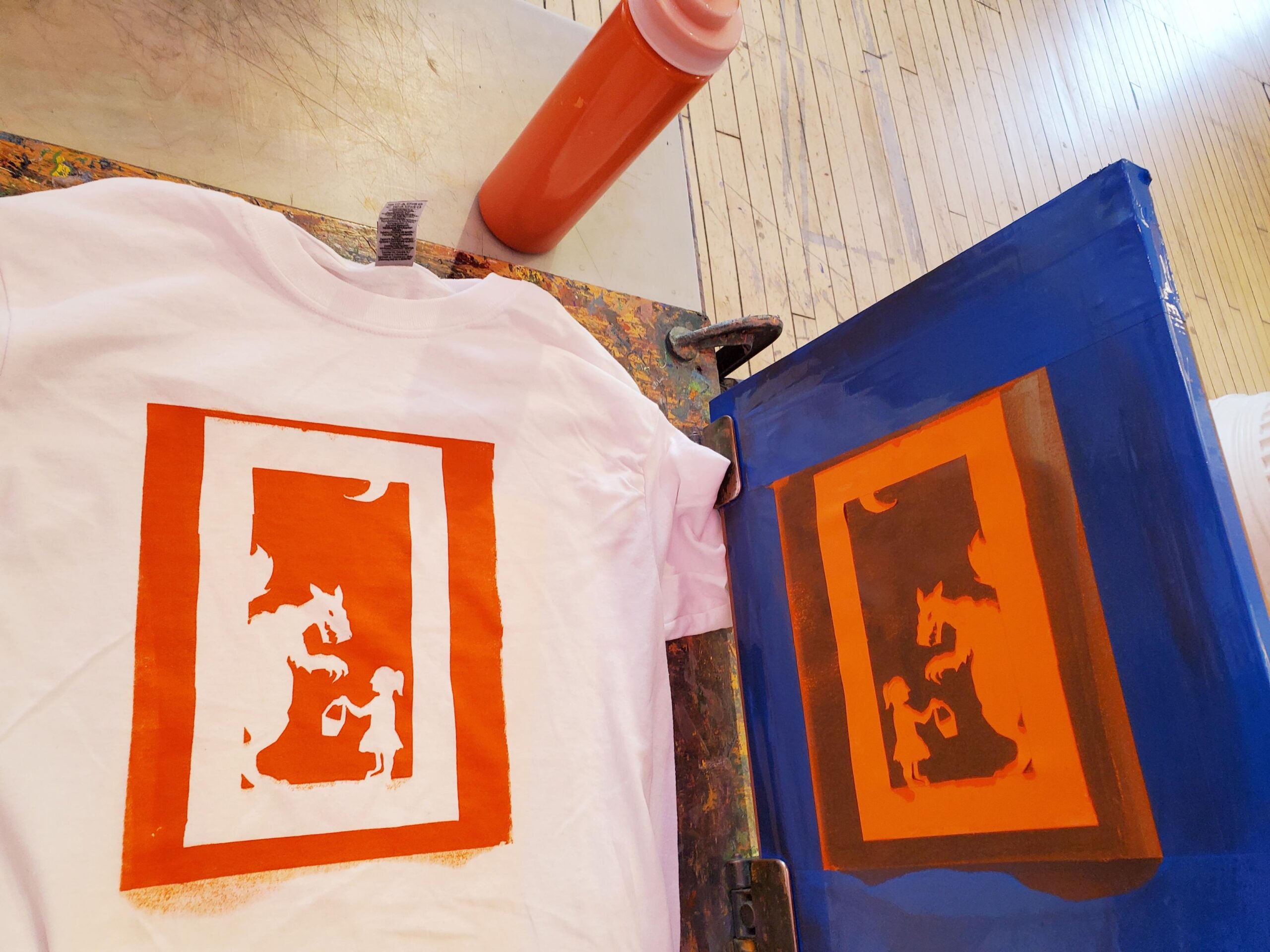 A t-shirt has just been screenprinted onto with orange ink. The screenprinted image shows a dragon and a child holding a basket.