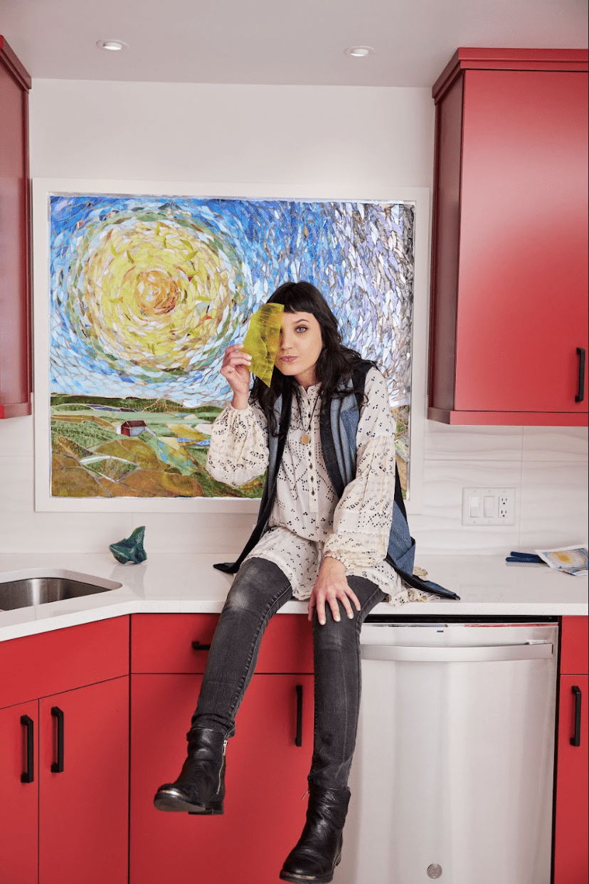 Artist Sarah Cohen sits on a kitchen counter in front of a painterly glass mosaic. Sarah is a white woman with long brown hair and she holds a piece of yellow glass in front of her right eye.