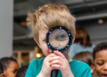 A child looks through a magnifying glass into the camera