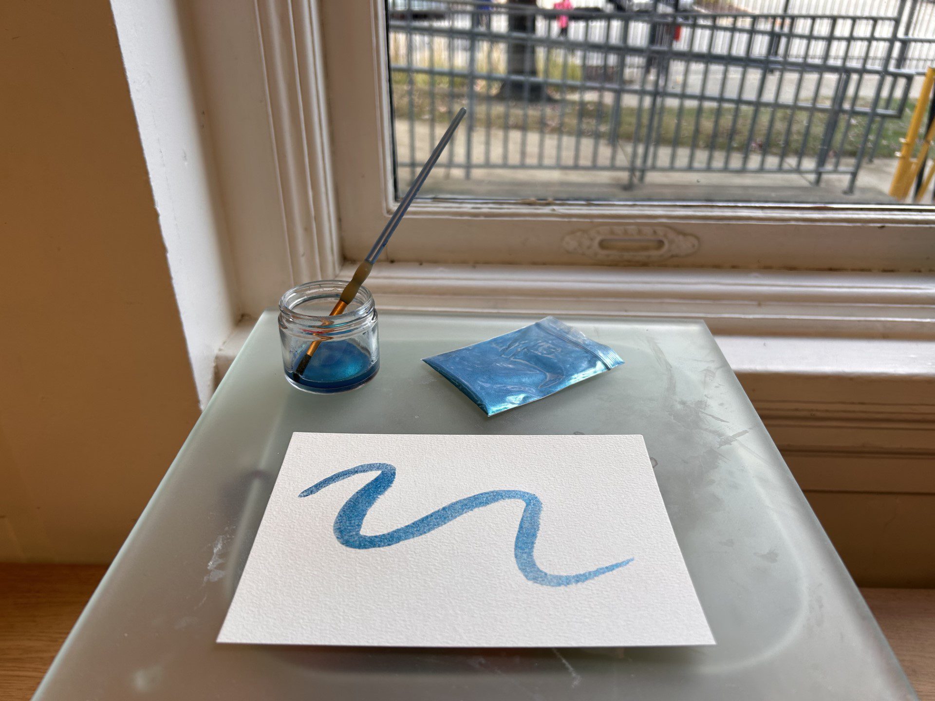 A piece of paper with a squiggly line of shimmery blue watercolor on it sites on a surface. Behind it are a small jar of the blue shimmery powder and blue shimmery liquid with a paintbrush in it.