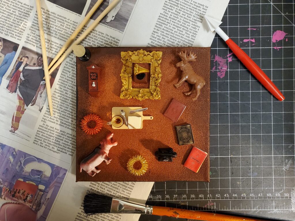 A brown painted canvas with small figurines and found objects glued on lays on a cutting mat