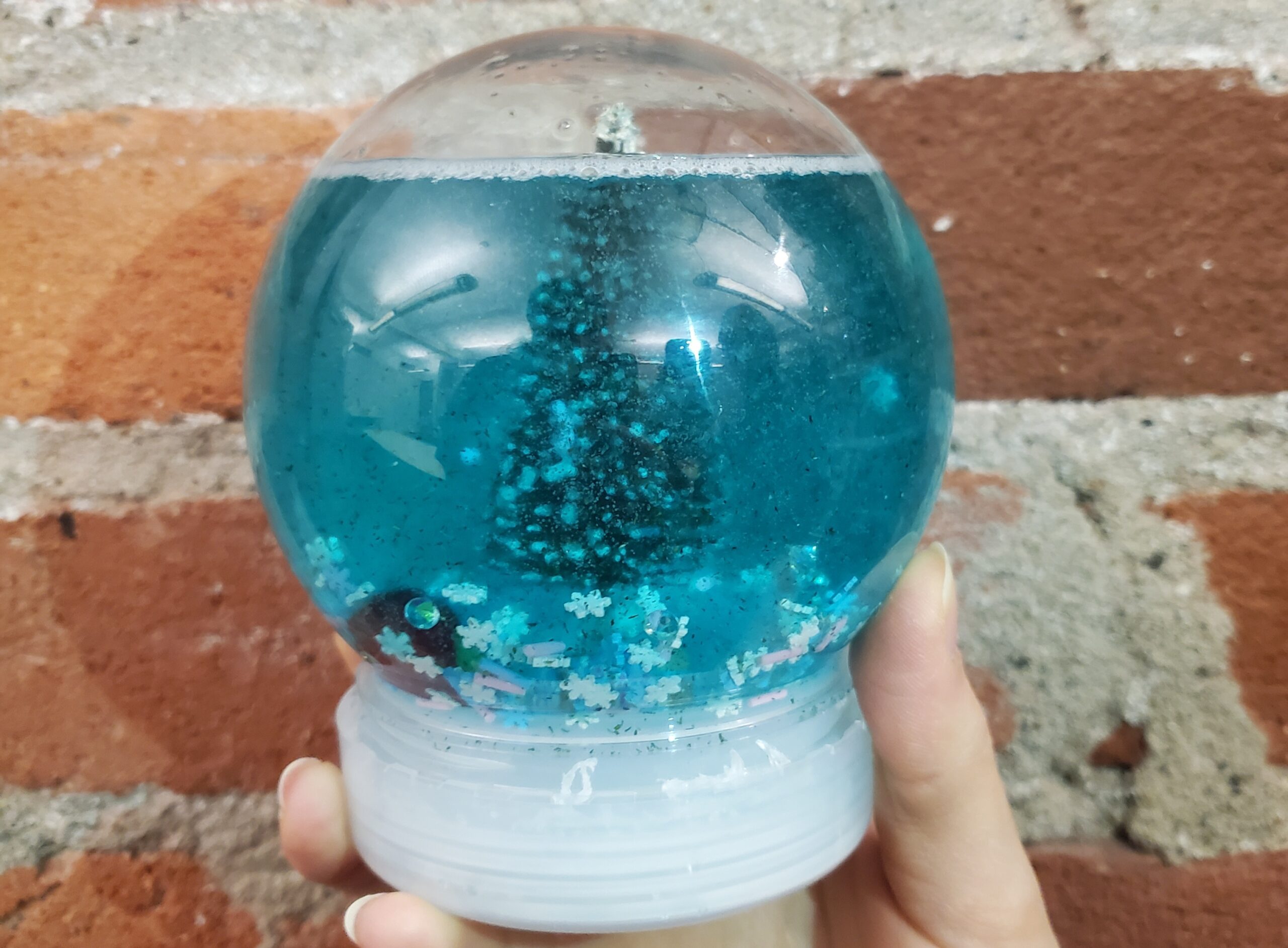 A hand holds a snow globe with an evergreen tree surrounded by blue water and snowflakes.