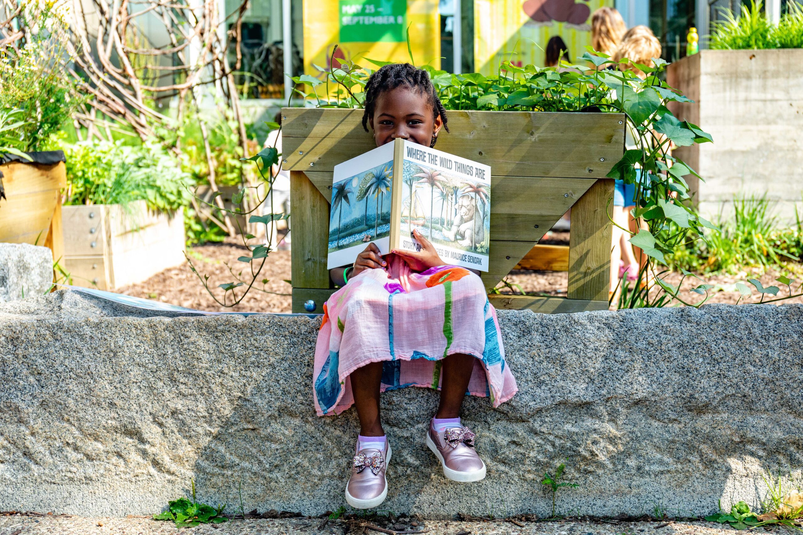 a girl sits outside holding the book "Where the Wild Things Are"